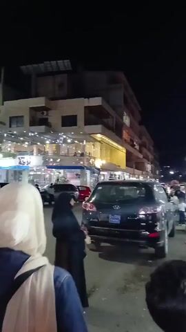 The video shows a woman wearing a white hijab and a beige jacket walking along a busy street at night. The street is lined with restaurants and shops, and there are many cars parked along the side of the road. The woman stops and takes a selfie with her phone, revealing her face. The phone has a light blue case with floral designs.