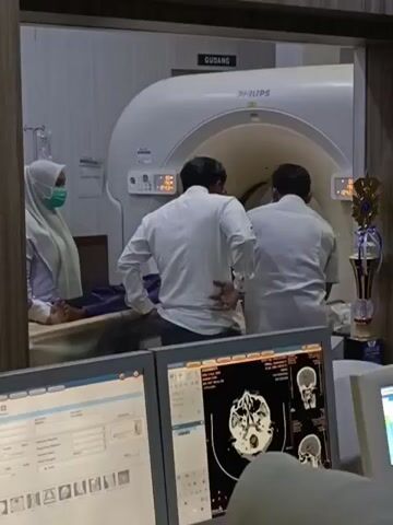 In a dimly lit medical room, a patient lies on a gurney beneath a large, white Philips CT scanner. The patient is partially obscured, but appears to be wearing a light-colored garment. Two medical professionals, both wearing white shirts, stand by the scanner, one with their back to the camera and the other facing the scanner. A third medical professional, wearing a hijab and a white uniform, stands to the left of the scanner, looking towards the patient. In the foreground, two computer monitors display medical images, including a detailed scan of a human brain. The room also contains a trophy on a stand to the right of the scanner, and a sign on the wall above the scanner reads "GUDANG," which is Indonesian for "Warehouse." The overall impression is of a medical diagnostic procedure in progress.