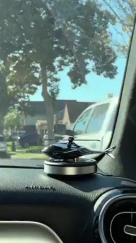 The video shows a small, black helicopter toy sitting on a silver disc on the dashboard of a car. The toy has rotating blades. The car is moving through a residential neighborhood on a sunny day. Houses and trees can be seen through the windshield. The dashboard is black and has a textured surface.  The video is shot from inside the car.