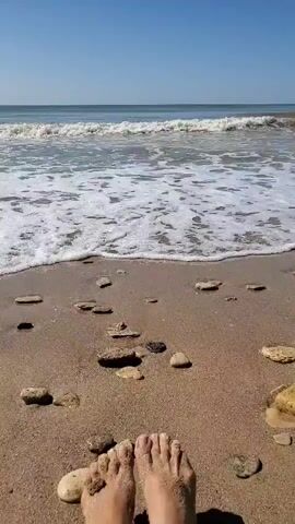 A first-person perspective video shows a pair of bare feet sunk into the sand on a beach. The camera is angled downwards towards the feet and the sandy shore. Gentle waves are rolling in from the ocean, creating white foam as they reach the shore. The ocean is a calm blue under a clear sky. Small rocks and pebbles are scattered on the sand around the feet. The scene is peaceful and depicts a moment of relaxation at the seaside.