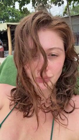 A close-up selfie shows a young woman with red, wavy hair, likely at a beach. She is wearing a green bikini top and a necklace. Her face is turned slightly, and she has freckles and a subtle smile. Her hair is partially obscuring her face, giving a windswept effect. In the background, there are glimpses of beach chairs and a rustic wooden structure, possibly a beach bar or restaurant. The lighting suggests it's daytime, and the environment appears to be a tropical beach setting. The woman's expression conveys a relaxed and happy mood.