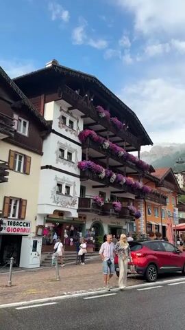 A couple is walking across a street in a European town. The woman is wearing a patterned top and wide-leg pants, while the man is wearing a striped shirt and shorts. They are walking towards a red SUV parked on the right side of the street. In the background, there are buildings with balconies adorned with flowers, shops, and mountains. The sky is partly cloudy.