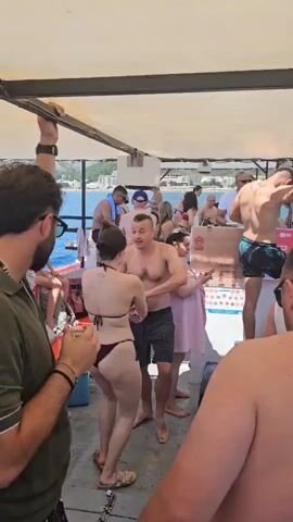 A group of people are on a boat party. A man in a green shirt is looking towards the left of the frame, holding a drink. To his right, a woman in a bikini is dancing with a man. Several other people are visible in the background, some are sitting, some are standing, and some are dancing. The boat is covered by a canopy. In the background, water and hills with buildings can be seen. The weather appears to be sunny.