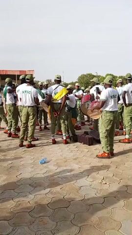 A group of individuals wearing matching khaki uniforms and green caps stand in a formation outdoors on a paved surface. Many of them are holding and playing drums. Some individuals have the letters "NYSC" printed on the back of their white t-shirts. The environment appears to be a sunny day with a light-colored sky. There are some water bottles on the ground. The individuals seem to be part of a band or performance group.