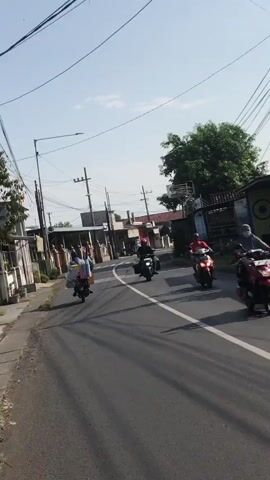 The media file captures a daytime scene on a paved road in Modong, Indonesia, under a clear sky. The sun is shining, casting shadows on the road. The road itself appears to be a two-lane street with a white dividing line. On the left side of the road, there is a sidewalk with some vegetation and buildings. On the right side, there are more buildings and trees.

The primary subjects are people riding motorcycles. In the foreground, a person wearing a red shirt and helmet is riding a motorcycle, heading away from the camera. Further down the road, another person wearing a helmet and dark clothing is riding a motorcycle, also moving away. In the distance, a third motorcycle is visible, carrying multiple people, one of whom is wearing a helmet. These individuals appear to be engaged in daily transportation.

The setting suggests a moderately urban or semi-urban environment, likely a residential or commercial street given the presence of buildings and utility poles with numerous wires. The architecture of the buildings appears to be consistent with Indonesian styles. The overall atmosphere is one of ordinary daily activity. There is no visible text in the media file.