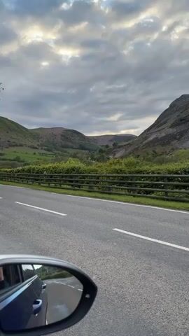 The video shows a view from inside a moving car, focusing on the road ahead and a landscape with mountains. The road is asphalt with white lane markings. A wooden fence separates the road from a grassy area with green bushes and trees. Beyond the fence, there is a valley between two mountains under a cloudy sky. The mountain on the right has a rocky, gray slope, with some vegetation. The left mountain appears greener with more vegetation cover. The view is partially framed by the car's side mirror in the lower left corner. The clouds are varied in color, ranging from light gray to darker shades, suggesting possible sunlight breaking through. Overall, the scene portrays a drive through a mountainous rural area on a cloudy day.