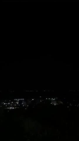 The video shows a dark, nighttime aerial view of a city. The lights of the city are visible in the distance, twinkling through the darkness. The camera appears to be panning slowly, offering a wide perspective of the urban landscape. The sky is completely black, suggesting it is a clear night with no moonlight. The overall impression is one of a serene, quiet night in a populated area.