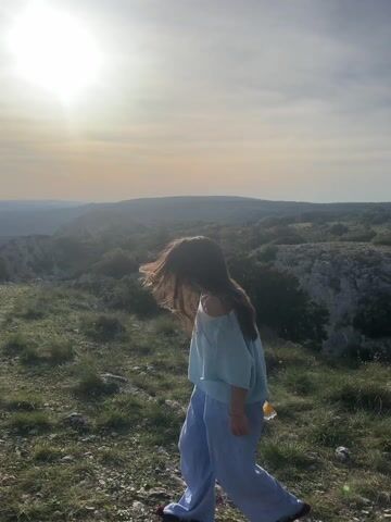 A woman with long, dark hair is walking on a grassy, rocky hillside. The sun is bright in the sky behind her, casting a warm glow. She is wearing a light blue off-the-shoulder top and loose, light blue pants. In her hand, she carries a translucent bottle. In the background, there is a landscape of rolling hills and trees. The sky is cloudy.