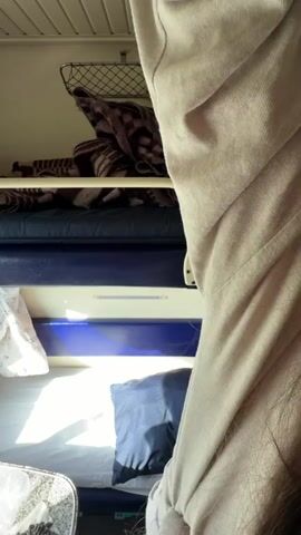 A woman with long brown and blonde hair is in a train compartment. She lowers a light-colored garment. She sits on the lower bunk of a bunk bed. The top bunk is covered with a striped blanket. She glances out the window. There's a small blue pillow on the bed beside her. Above the bed, there's a wire rack.