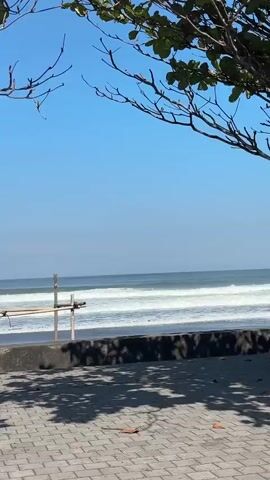 A vertical video shows a sunny beach with blue skies and calm ocean waves. In the foreground, a walkway with patterned paving stones is visible, covered in dappled shadows from overhead foliage. A low concrete barrier separates the walkway from the dark sand of the beach. Beyond the beach, gentle waves break on the shore, with the ocean stretching to the horizon under a clear blue sky. A simple wooden structure, possibly scaffolding, stands on the beach near the water's edge. The camera is stationary and the video pans slightly to the right.