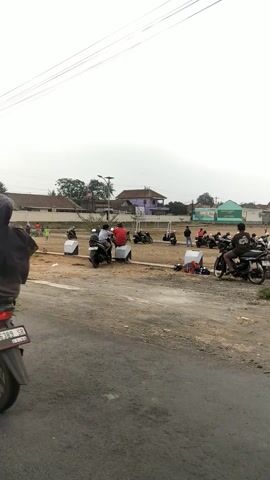 The video pans across a dusty, outdoor scene with a gray asphalt road in the foreground. In the background, there is a large, open dirt area with several motorcycles and a few people. Some of the motorcycles are parked, while others appear to have riders on them. There are also some small white boxes scattered around the area. In the distance, there are several buildings, including a purple house and a green building that looks like a school or community center. The sky is overcast. One of the motorcycles has a person wearing a red shirt on it. A black car with colorful stickers is parked in the foreground. Another silver car is parked on the side of the road.
