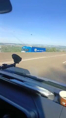 The video is filmed from the interior of a vehicle, looking out through the windshield. It appears to be daytime with a clear blue sky and bright sunlight casting shadows. The vehicle is moving along a highway, indicated by the road markings and the view of the landscape blurring past. On the right side of the road, there is a concrete barrier. Beyond the barrier, there is a green, wooded hillside with some buildings visible in the distance. A large blue and white billboard with text "JANSSEN WWW.CTS-ONLINE.COM" is partially visible on the left side of the road. On the dashboard of the vehicle, a black smartphone is mounted in a holder and a red beverage can is in a cup holder. The perspective suggests the viewer is a passenger or driver.