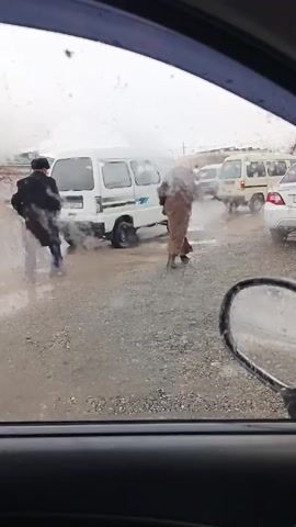 The view from inside a car looking out at a gravel lot on a gray, wet day. Rain or sleet streaks the window. A white minivan is visible in the middle of the frame, slightly obscured by steam or smoke rising from its rear. Two individuals are walking near the van: a person in a dark coat and hat, and another in a long brown coat and a light-colored head covering. They appear to be walking away from the van, which is partially stuck in a puddle. Other parked cars are visible in the background, including a white sedan to the right. The car's side mirror is visible in the foreground.