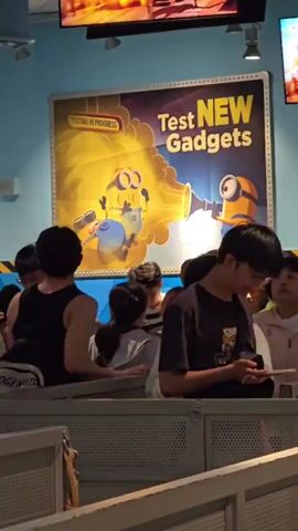 The video shows a line of people waiting in what appears to be a themed attraction or queue area. In the background, a large poster features two Minion characters from the Despicable Me franchise. The poster has the text "Test NEW Gadgets" and "TESTING IN PROGRESS" written on it, with one Minion holding a device that is emitting a yellow beam of light towards the other Minion. The people in the foreground are diverse in age and gender, and most are looking down at their phones or ahead at the line. One person in the foreground is wearing a white jacket, another a green t-shirt, and one a black tank top. The environment seems to be indoors, with blue walls and a metal railing forming the queue barrier. The lighting is moderate, and the overall atmosphere appears to be casual, with people waiting patiently.