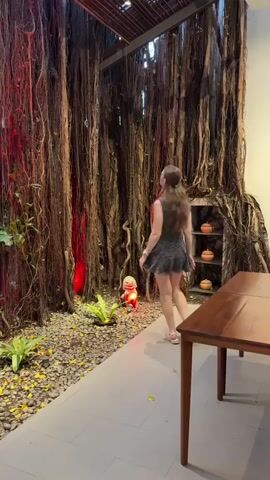 A young woman in a black dress and sandals walks through an indoor space with a rustic, nature-inspired design. She carries a small purse and pauses to look around. The environment features a large display of hanging tree roots, gravel, plants, and decorative pots, with a wooden table in the foreground. Red lighting illuminates some of the roots, creating a dramatic effect.