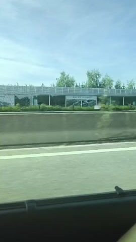 Here is a description of the video:

The video appears to be shot from a car traveling on a highway. The camera is pointed towards the right side of the vehicle, offering a view of the roadside scenery. Initially, the camera captures an Audi dealership, displaying large banners advertising different Audi models, including the Audi RS e-tron GT and the Audi Q4 Sportback e-tron. The dealership is characterized by a modern, gray-toned architecture, with greenery partially obscuring the view.

As the car moves forward, the camera then pans across the dealership’s main building, identifiable by the Audi logo prominently displayed on the rooftop. The building also features a Pirelli logo. A concrete barrier separates the highway from the dealership property.

The video then continues to show a white van traveling on the highway next to the filming vehicle. The view is brief, focusing on the side of the van as it passes. The interior of the filming vehicle is partially visible, as a dark piece of the car's interior is seen in the foreground.