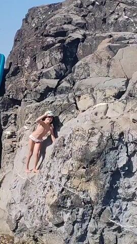 A young person wearing a bucket hat and bikini bottom is climbing on a rocky surface. The person is facing away from the camera and is using their hands and feet to ascend the rocks. The environment appears to be outdoors with a clear sky. A blue inflatable object is partially visible on the left side of the frame.