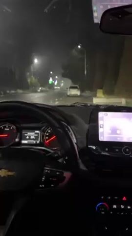 The video shows a dashboard view from inside a Chevrolet car driving at night on a wet road. Streetlights illuminate the road ahead, and another car is visible in the distance. The windshield wipers are active. The car's dashboard is visible, showing the speedometer, fuel gauge, and a digital display. The central console includes an infotainment system and climate control buttons. The view is primarily dark with illuminated elements from the car's dashboard and external lights.