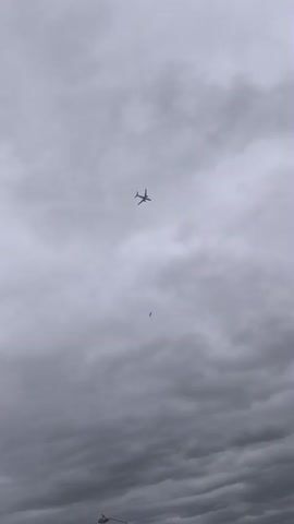 The video displays a view of a cloudy sky with an airplane flying high above. There is also a smaller object, possibly a bird, in the sky beneath the airplane. The scene appears to be set outdoors, as indicated by the sky and the faint outlines of buildings and possibly a lamppost at the bottom of the frame. The weather is overcast and gray, suggesting a dull or possibly rainy day. The time of day is not explicitly clear, but the lighting suggests it is daytime. There are no people clearly visible in the foreground or the sky. The overall mood is somewhat somber due to the cloudy conditions. No text is visible in the video.