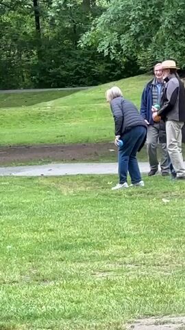 This is a slow-motion video taken outdoors on a grassy field. In the foreground, a woman with blonde hair wearing a grey puffer jacket and blue pants is bending down, holding a blue ball. Behind her, three people are standing; one man in a blue jacket and glasses, a man in a grey jacket and hat, and a third person who is partially obscured. In the background, there are trees and a stone wall. The video captures the woman's throwing motion. The scene transitions to a blurry image of a man in a denim jacket walking away from the camera, followed by shots of trees and picnic tables. The video then returns to focus on a woman wearing a light-colored sweater and a patterned skirt, who appears to be throwing a yellow ball.
