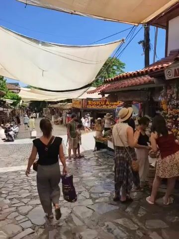 The video shows a busy outdoor marketplace on a sunny day. A cobblestone path winds through the market, lined with stalls selling various goods. In the foreground, a woman with dark hair walks away from the camera, carrying a purple shopping bag. She is wearing a black top and grey pants. To her right, a man in a grey shirt and shorts stands with a child. Further down the path, several other people are browsing the stalls. The stalls themselves are covered by white canopies, providing shade from the sun. One stall, labeled "TOSTCU AHMED," appears to be selling food. Another stall showcases an array of ice cream cones and what looks like a selection of ice cream in a refrigerated display case. A sign on this stall reads "DONDURMA," which means ice cream in Turkish. The overall atmosphere is lively and bustling, with shoppers and vendors engaged in commerce. The architecture of the buildings lining the market appears to be traditional, with red-tiled roofs. The sky is clear and blue, indicating good weather.