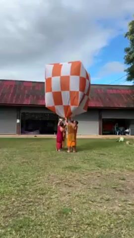 Four people are gathered in a grassy yard, holding up a large, cube-shaped object. The object is covered in an orange and white checkerboard pattern. The people are wearing robes, three in a light orange color and one in a deeper red. Behind them is a long, one-story building with a red corrugated metal roof and open garage doors. The sky is partly cloudy with patches of blue. The people appear to be preparing to release the checkerboard object into the air.