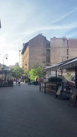 The video shows a street scene in a European city. On the left side, there are several outdoor seating areas for cafes or restaurants, with tables and chairs. A few people are seated at these tables. Further down the street, there are more cafes with awnings and seating.

In the center of the street, several people are walking in both directions. There are also some electric scooters parked along the side of the street.

The buildings lining the street are multi-story structures with brick and stone facades, typical of older European architecture. Some of the buildings have balconies or window boxes with plants. Trees are planted along the street, providing some shade.

The sky is blue with some scattered clouds, suggesting it is daytime and likely pleasant weather. The overall impression is that of a lively, pedestrian-friendly street with outdoor dining and a relaxed atmosphere.