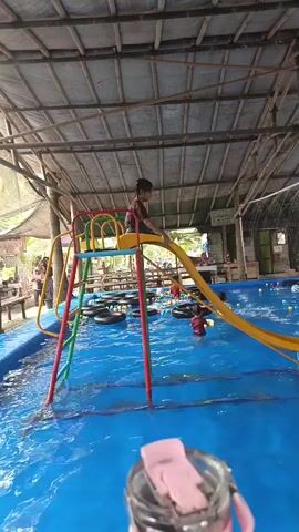 A young child slides down a yellow playground slide into a blue swimming pool. The pool is covered by a thatched roof structure. There are various inflatable rings floating in the water and colorful balls scattered around the pool. The child splashes into the water and then stands up, smiling at the camera. Another person is visible in the background, also in the pool. The scene appears to be a recreational area.