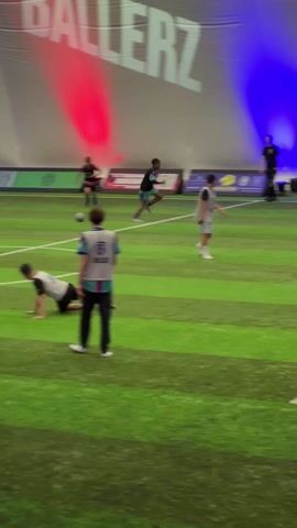 The video shows an indoor soccer game. Several young players are on the green and white striped field. One player wearing a black and red jersey dribbles the soccer ball. Other players are walking and running. A referee stands on the sideline. A large sign reading "BALLERZ" is visible in the background with red and blue lights shining behind it. There are advertisements and sponsor logos along the wall.