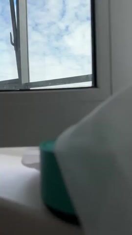 The video shows a view of a white window with blue sky and clouds visible through the open section. In the foreground, slightly out of focus, is a green container with a white tissue or cloth on top. The scene is indoors, with the window on a white surface, possibly a windowsill.