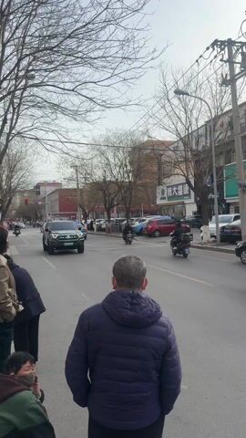 The video shows a street scene in China. A gray SUV drives along a city street lined with buildings. People are on the sidewalks, and other vehicles are driving in the street, including cars, trucks, and motorcycles. The buildings have signs in Chinese characters. The sky is overcast. The SUV has a spare tire mounted on the back.