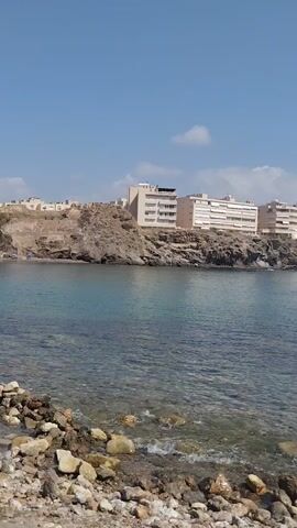 The video shows a coastline with buildings on a rocky cliff overlooking a body of water. The water is clear and shows rocks on the seabed. In the distance, there is a bridge or pier structure extending over the water, with more buildings visible beyond it. The sky is blue with some scattered clouds. The foreground is a rocky beach.