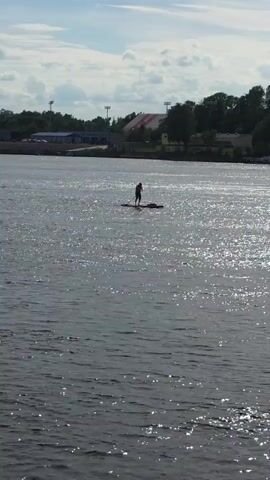 The video captures a river scene on a bright, partially cloudy day. A person on a paddleboard is visible in the distance, paddling in the sun-dappled water. The camera pans right, revealing a large concrete bridge stretching across the river. The camera then pans further right, showing an urban environment on the riverbank with various buildings, including a large industrial-looking structure with a tall smokestack. Two people are seen kayaking on the river, paddling in unison. The water is a dark, shimmering blue, reflecting the sunlight.