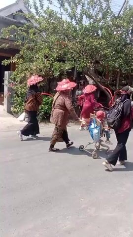 The video shows a street scene with several people and vehicles. In the foreground, three people are walking, one of them pushing a stroller. They are all wearing hats. Behind them, a person is riding a motorcycle. Further down the street, another person is riding a motorcycle with a large box on the back. In the background, a decorated vehicle is visible. There are houses and trees lining the street. A flag is also visible on a flagpole. The scene appears to be outdoors on a sunny day.