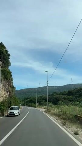 The video shows a drive on a two-lane road in a natural setting. The road is paved with a white line dividing the lanes. On the left side of the road, there is a rocky cliff overgrown with greenery. On the right, a hill covered with trees is dotted with multiple wind turbines on its peak.  Street lights line the side of the road, and there's a 60 speed limit sign. Cars, including a truck, are driving on the road.  The sky is partly cloudy with patches of blue.