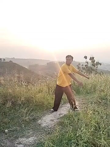 A single man stands on a dirt path in a grassy, hilly landscape, possibly at sunrise or sunset. He is wearing a yellow short-sleeved shirt and dark pants. The man performs gentle, flowing movements with his arms and upper body, resembling stretching or a light dance. He looks up and smiles, appearing happy and relaxed. The background features distant hills and a bright, low sun creating a glare. The surrounding area is covered in tall, sparse grass and some small trees or bushes. The setting is natural and outdoor, with clear skies. No text is visible.