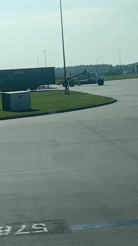 This is a video clip filmed from a slightly elevated perspective, likely from a vehicle or building. The scene is an outdoor paved area, resembling a parking lot or staging area, with a clear blue sky and bright sunlight indicating it's daytime. The weather appears to be clear and sunny.

In the foreground, a portion of a dark asphalt surface is visible, marked with white and blue painted lines, including what looks like a parking space number, "579". Further back, a bright green patch of grass forms a curved island. On this grass, a dark shipping container with the "prime" logo is parked. Behind the shipping container, a utility box with the number "11" on it is visible.

Dominating the mid-ground is a light-colored tow truck with its boom extended. The tow truck is positioned on the edge of the paved area, near the grass, and appears to be either parked or maneuvering. A street lamp stands tall between the shipping container and the tow truck, its pole casting a faint shadow. In the distance, more parked containers and a line of trees are visible, suggesting the area might be a logistics hub or distribution center. The overall atmosphere is industrial and functional. No people are clearly visible, but the presence of the tow truck implies ongoing operations. There is a speed limit sign with "30" visible near the tow truck.