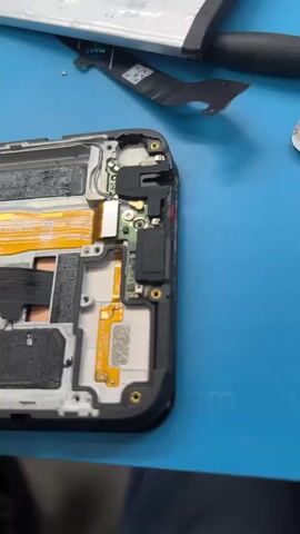 The video shows a disassembled smartphone, revealing its internal components. The motherboard is visible, along with various ribbon cables, connectors, and what appears to be a camera module. Some screws and a SIM card tray are scattered on a blue mat, suggesting a repair or disassembly process. A portion of a white battery is also present in the upper right corner. The scene is likely a workspace for electronics repair.