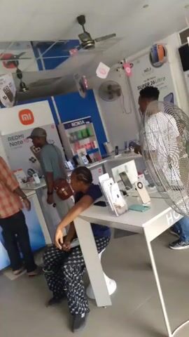 The video shows the interior of a cell phone store. A man in a red and black plaid shirt stands on the left and turns to look at someone off-screen. Another man wearing a gray shirt and carrying a brown bag is looking at phones on display. A woman with braided hair is seated at a white table, examining a piece of paper. A person wearing a white patterned shirt stands to the right, near a large white fan. A display for Redmi Note 15 is visible on the left. Another display case for Nokia phones is in the background. There are ceiling fans and promotional signs hanging from the ceiling. The scene is well-lit.