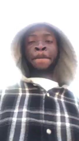 A man is walking outside in a bright, sunny environment. He has dark skin, a beard, and is wearing a black and white plaid shirt with a grey hood. His hood is up. In the background, there is a road with cars. He is looking directly at the camera and appears to be filming himself.