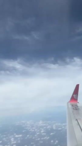 The video provides an aerial view from an airplane window, looking out over a vast expanse of clouds. In the foreground on the right side, a portion of the airplane's wing is visible. The wingtip is red and features a white logo resembling a lion's head, characteristic of Lion Air or its subsidiaries. Below the red wingtip, some text is partially visible, which appears to be "HS-LJC" or similar, indicating a Thai-registered aircraft.

Below the aircraft, a thick blanket of fluffy, white cumulus clouds stretches out to the horizon, creating a soft, uneven surface. Above the aircraft, the sky transitions from a lighter, hazy blue near the horizon to a darker, more prominent greyish-blue with scattered, wispy clouds higher up. The overall impression is one of peaceful flight above the clouds during what appears to be daytime. The camera pans slightly downwards at one point, revealing a small part of the engine nacelle at the bottom of the frame, before panning back up to the horizon. The setting is likely above Thailand, given the aircraft registration and brand.