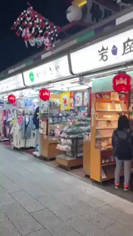 The video shows a street scene at night, lined with shops under awnings with bright white signage. Red lanterns and decorative white and pink floral arrangements hang above the street. Pedestrians walk along the paved path, with a building visible at the end of the street. The shops display a variety of goods, including clothing, souvenirs, and jewelry. People are dressed in warm clothing suitable for the night weather.