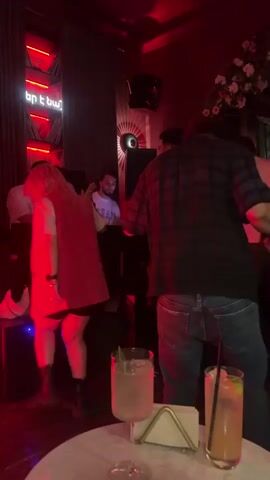 The video shows a dimly lit nightclub with red lighting. In the foreground, two cocktails are on a table. In the background, a DJ is seen behind a console, and several people are dancing. One person in a red vest is dancing, while another person in a plaid shirt is also visible. The overall atmosphere appears to be lively and energetic.