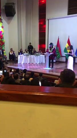 The scene shows a person giving a speech at a podium on a stage, addressing an audience.

The speaker is wearing a dark hat, a maroon-colored garment, and athletic shoes. They are standing behind a white podium with a document or notes on top. A floral garland is around their neck.

Behind the speaker, a table is covered with a white tablecloth and decorated with floral arrangements. Several people are seated behind the table. To the right of the speaker are two flags; one with green, yellow, and red stripes and one with a square red, yellow, and green pattern. To the speaker's left are white curtains and a stack of balloons.

The audience sits in rows in front of the stage, watching and recording the event with their phones. The room appears to be a large auditorium with wooden accents. Red lights shine in the top right corner of the stage.