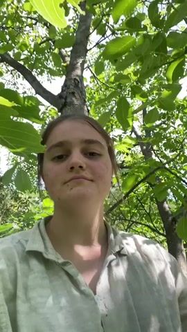 A young woman with brown hair, wearing a light-colored, striped shirt, is filmed in a wooded area. The camera angle shifts from her face to a view of tree branches and green foliage. Her bare feet are visible as she sits on a tree branch, looking down at the ground below. The scene appears to be outdoors during the daytime.
