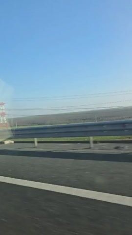 The video, taken from inside a moving vehicle, shows a view of a highway with a guardrail on the side. Beyond the guardrail is a field, and further in the distance, several electrical towers. The towers are painted white and red. A billboard with "IMPLANTODENT" written on it is also visible. A silver coupe passes by on the highway. The sky is a clear, bright blue. The video appears to be shot during the daytime.