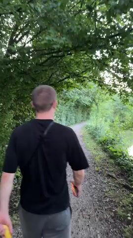 A man walks away from the camera down a gravel path lined with trees and foliage. He is wearing a black t-shirt and grey shorts. The path curves to the right, leading towards a body of water visible on the right side of the frame. The water reflects the trees and the sky. The overall scene suggests a natural, outdoor environment, possibly a park or nature trail, during daylight hours.