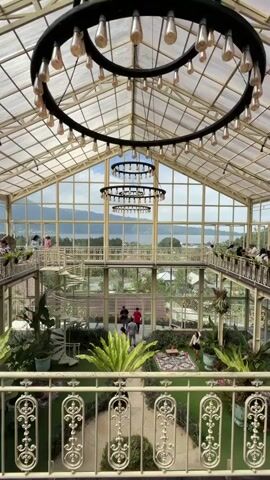 This video captures an interior view of a large, multi-level greenhouse or conservatory. The structure features a high, sloped glass ceiling and walls, allowing ample natural light and offering a scenic view of a body of water and mountains in the distance. Three large circular chandeliers with exposed bulbs hang from the ceiling. The interior is designed with lush greenery, including potted plants and a carpeted floor in some areas.

On the ground level, a pathway winds through artificial turf. Several people are visible: a couple in formal attire, possibly for a photoshoot, walks along the path; other individuals are scattered throughout the space, some appearing to be patrons or staff. A spiral staircase is prominent on the left side.

Multiple balconies or walkways are present on the upper levels, also lined with railings and people observing the scene below. The overall impression is one of a spacious, elegantly decorated venue with a focus on natural elements and an expansive view.
