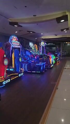 The video shows a row of brightly lit arcade games. The camera pans from left to right, focusing on the games. The first game on the left is red and blue, and appears to be a driving game. The next game is a Transformers-themed game with a large display featuring Megatron. This game is predominantly purple and blue, with flashing lights. The camera then moves to show more arcade games in the background, also lit with colorful lights. The overall environment is that of an arcade, with a dark ceiling and wooden floor. No people are visible in the video.