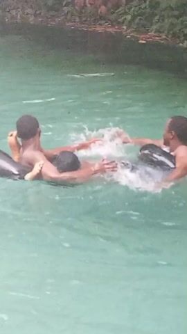 Two young men are shown floating in black inner tubes in a river of pale blue-green water. The man on the left is facing away from the camera. The man on the right is facing toward the camera, smiling and splashing with his arms. The water appears to be moving slowly, and there are trees and plants visible in the blurred background.
